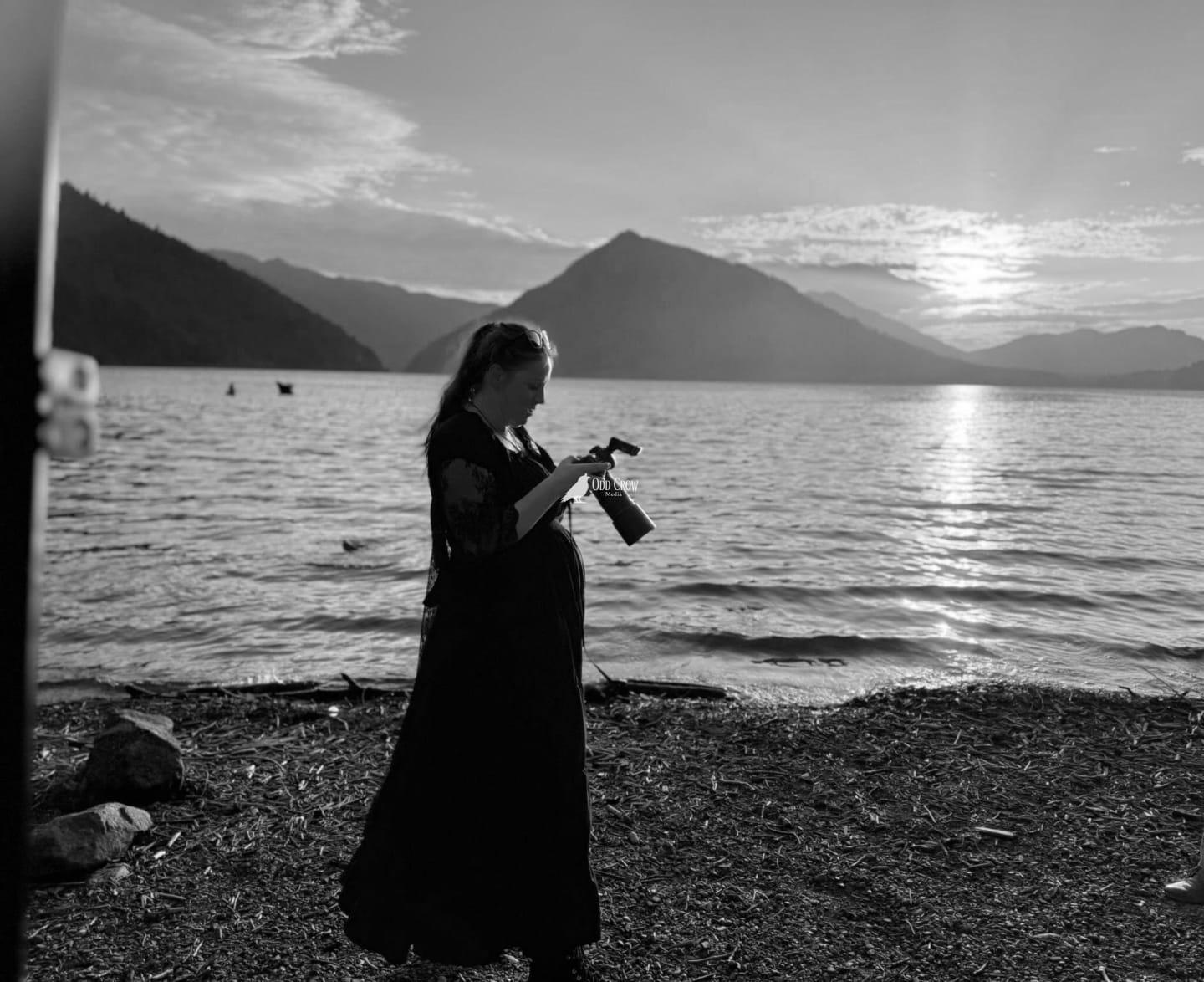 Black and white image of woman in flowing dress photographing on a beach with mountains and water in background