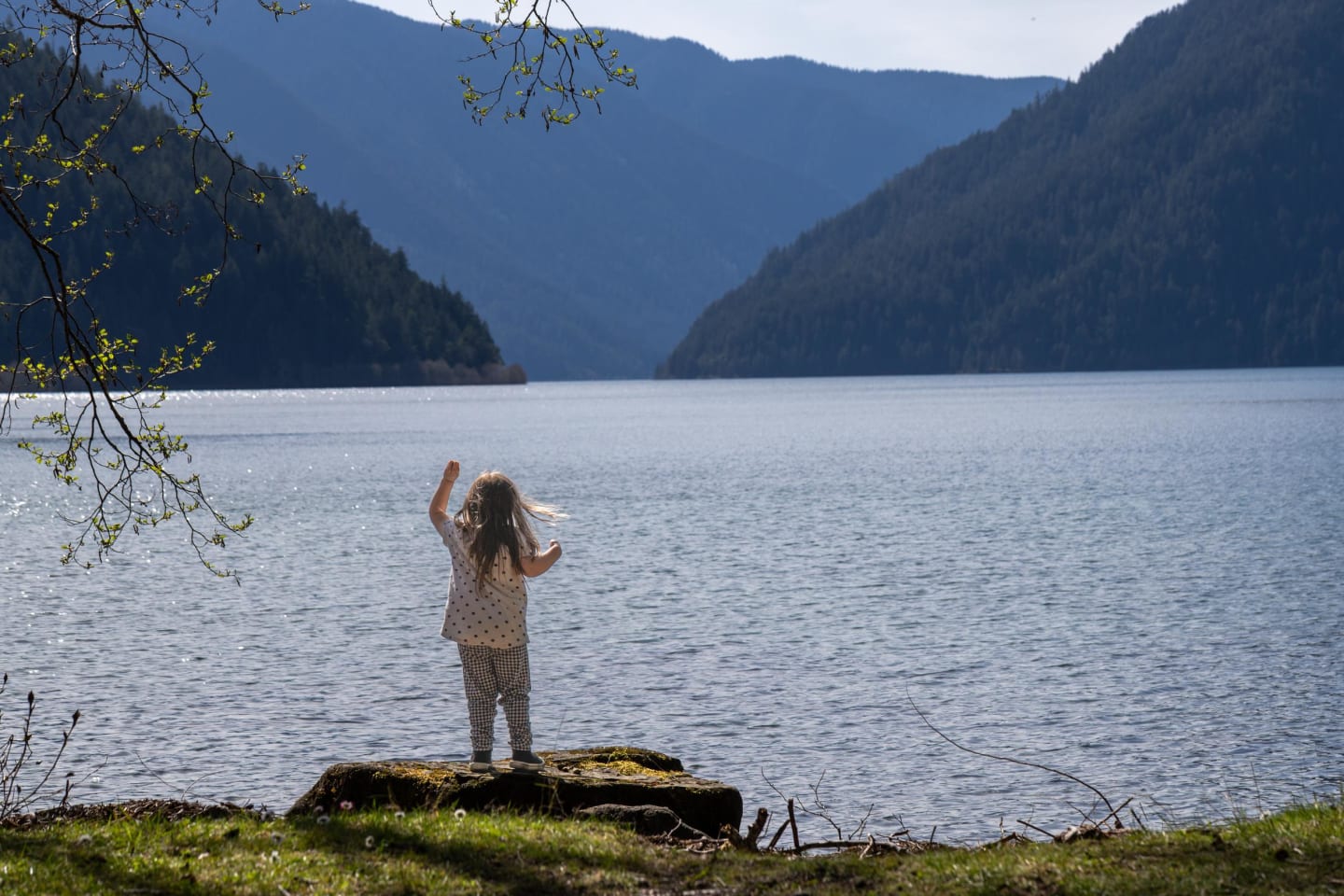 Young child with blonde hair waving from a moss-covered rock at the lake's edge, mountains and water stretching behind
