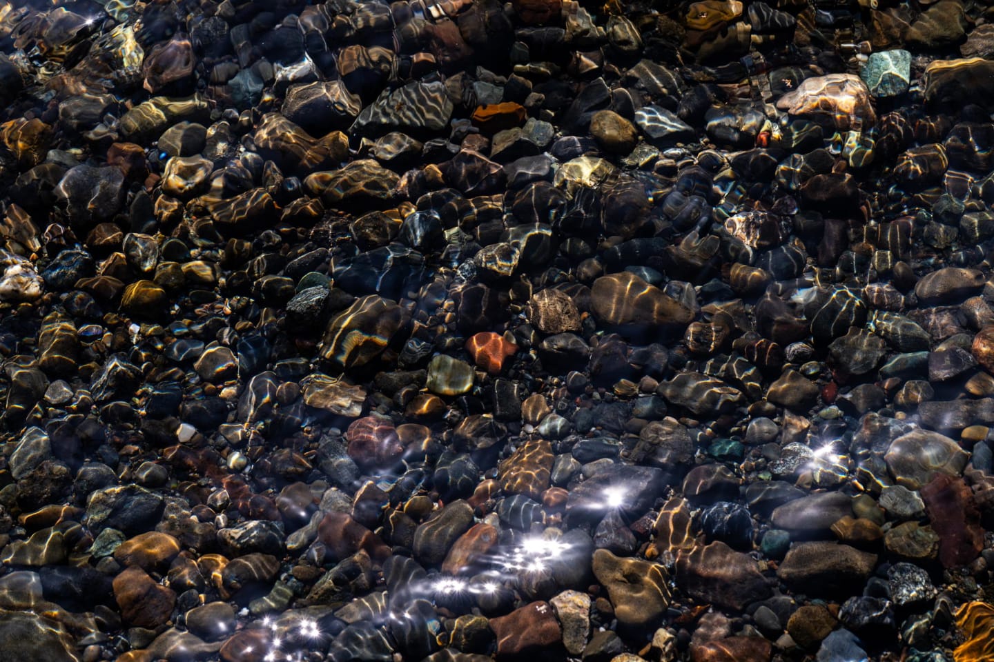 Close-up view of colorful stones beneath crystal clear lake water with sunlight creating sparkles on the surface