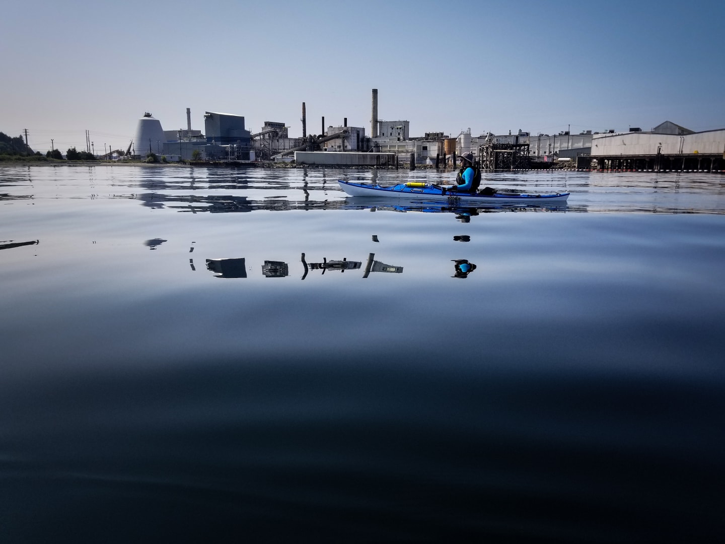 Kayaker paddling past industrial mill complex reflected in calm water