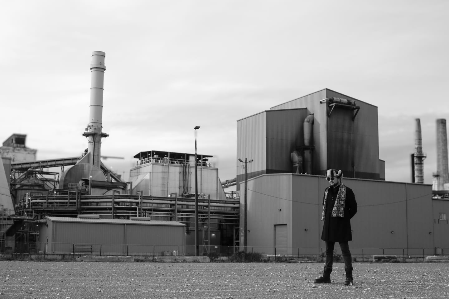 Person in dark clothing walking across industrial lot with mill structures towering behind