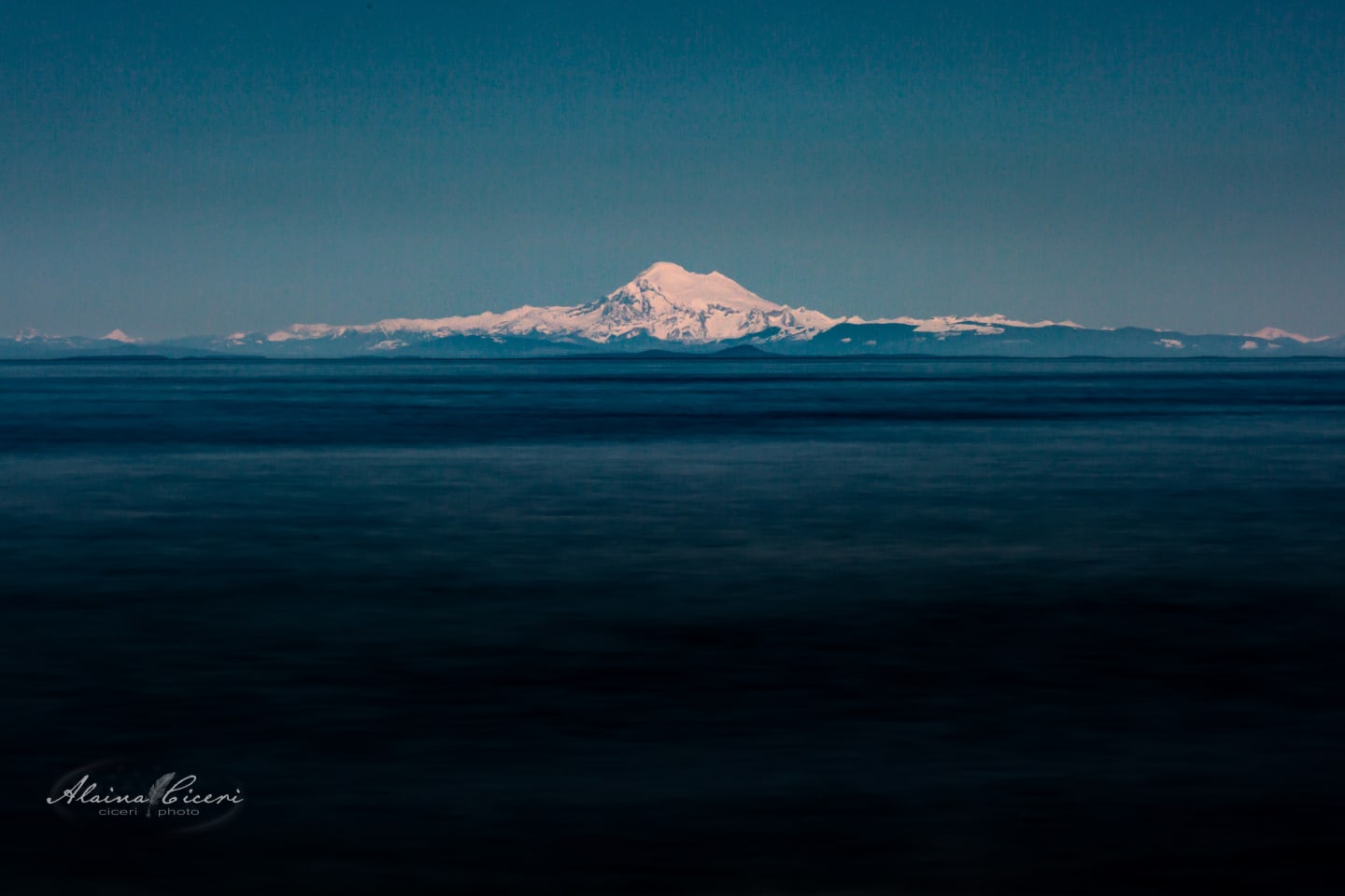 Black and white image of calm water with Mount Baker visible as a pale silhouette on the distant horizon