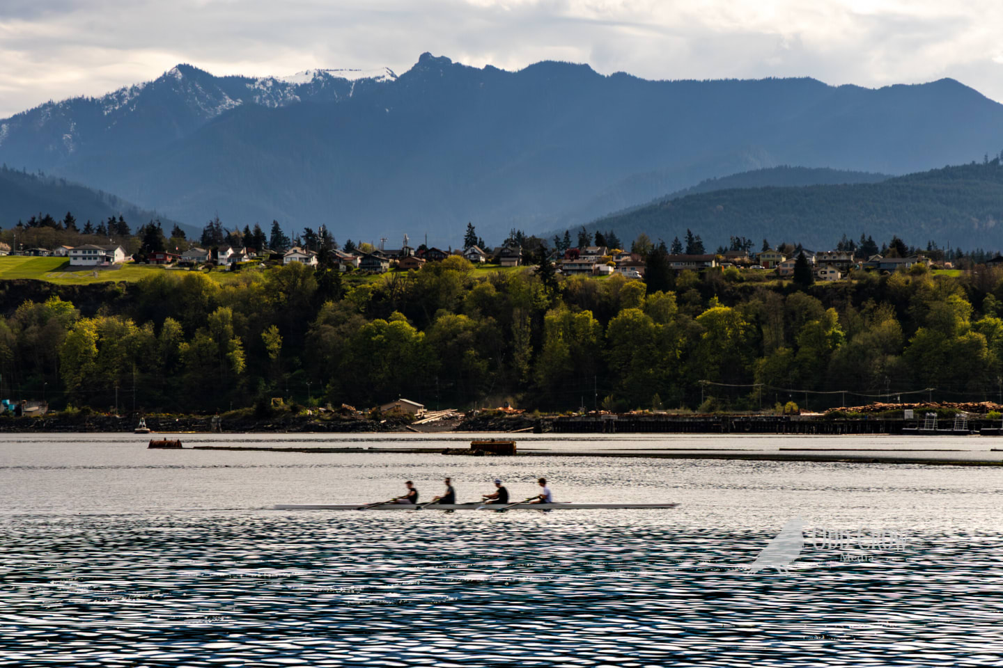 Three kayakers spread across calm harbor water with layered Olympic Mountains and Port Angeles residential areas in background