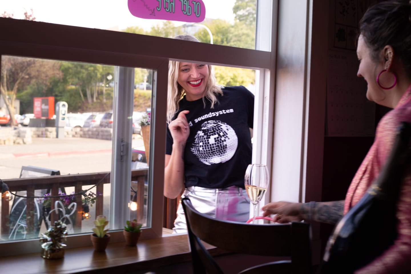 Woman in graphic tee laughing while holding wine glass, sitting by large windows with natural light streaming in