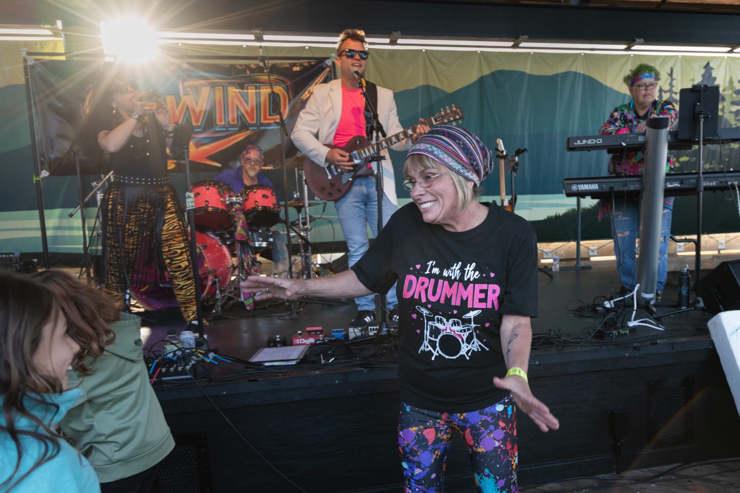 Woman in 'I'm with the Drummer' shirt dancing with a child near the front of the stage at Concerts on the Pier, full band visible performing behind them in late afternoon light