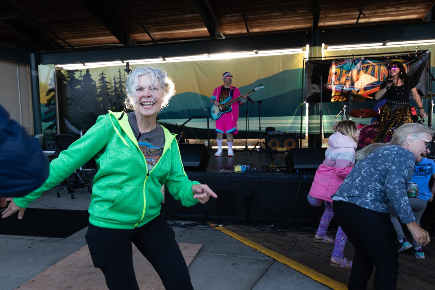 Older woman in bright green zip-up hoodie dancing joyfully at the front of the crowd at Concerts on the Pier, with the full Rewind band performing on stage in the background