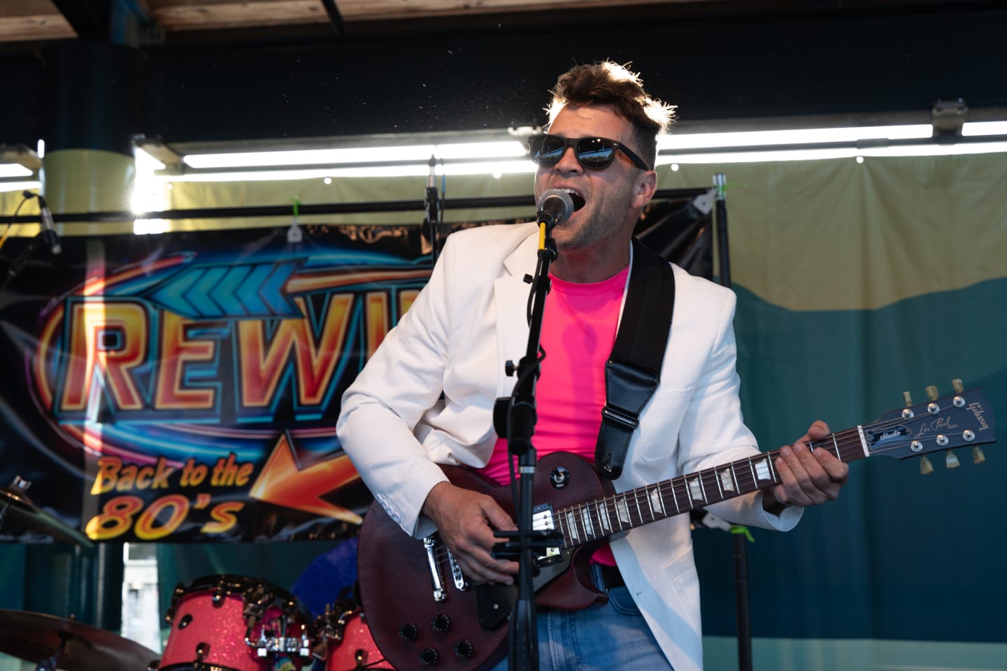 Guitarist and vocalist of Rewind in white blazer and pink shirt singing into a mic while playing red Gibson Les Paul guitar, Rewind Back to the 80s banner behind him