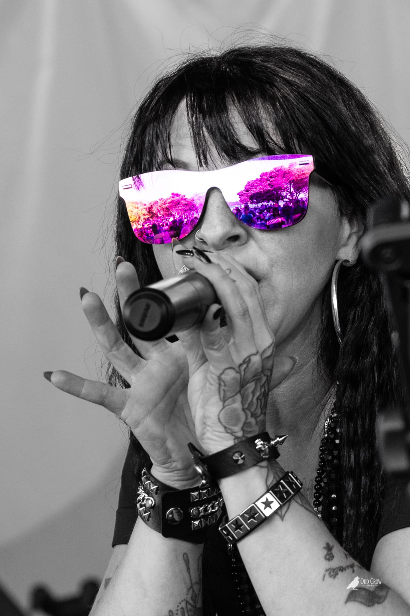 Close-up black and white portrait of the female lead singer of Rewind performing, pink mirrored sunglasses reflecting the crowd, microphone at her lips, tattoos visible on her hands