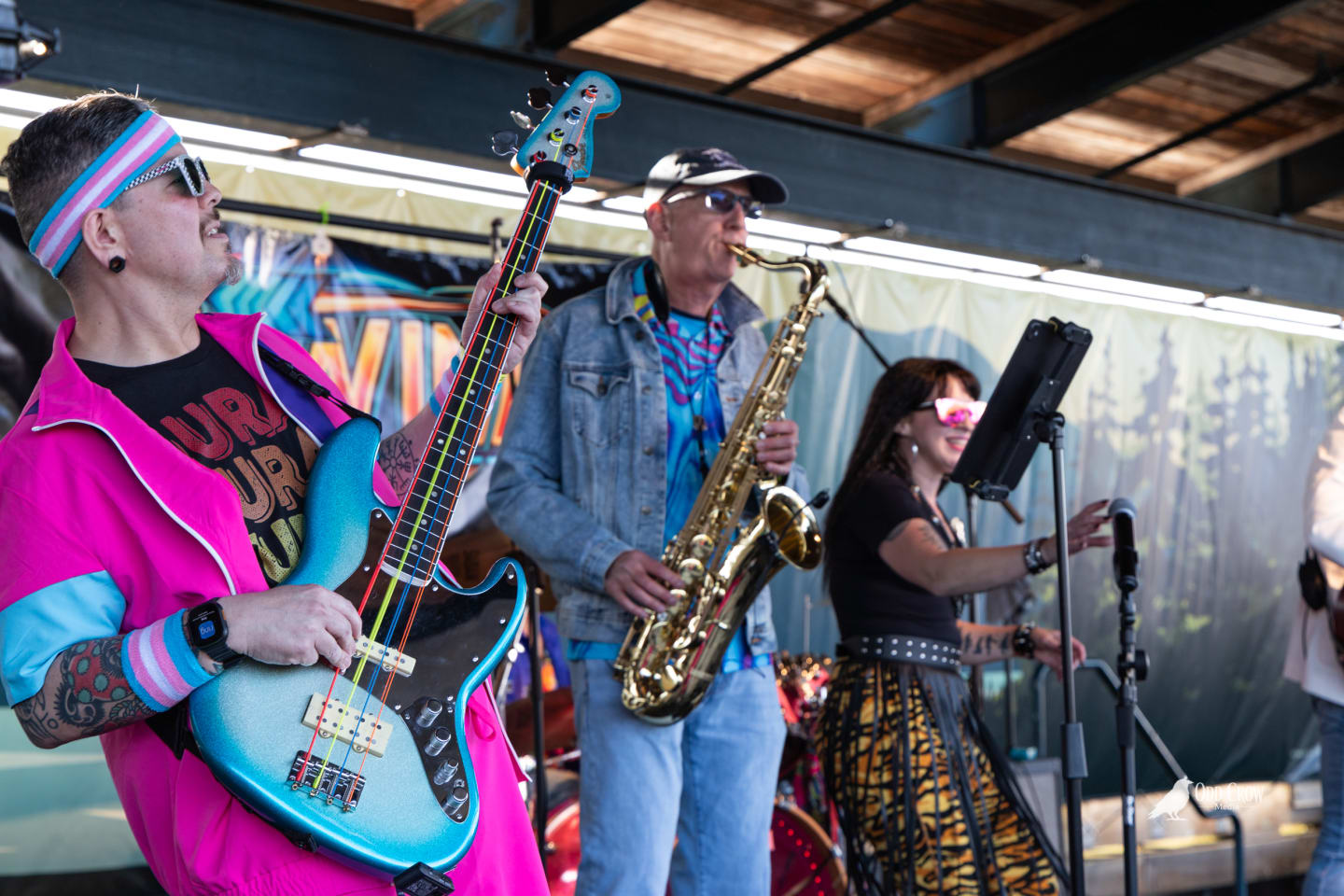Bass guitarist in neon pink tracksuit and striped headband alongside the saxophonist in a denim jacket, both performing on the Rewind stage at Port Angeles City Pier