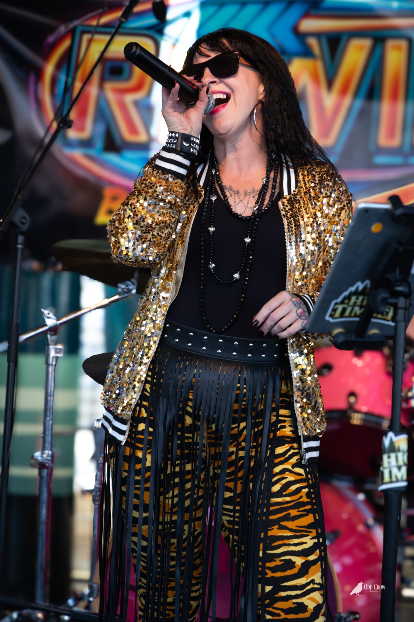 Lead singer of Rewind in gold sequin jacket and tiger print fringe skirt singing into a microphone, laughing mid-performance against the colorful Rewind banner