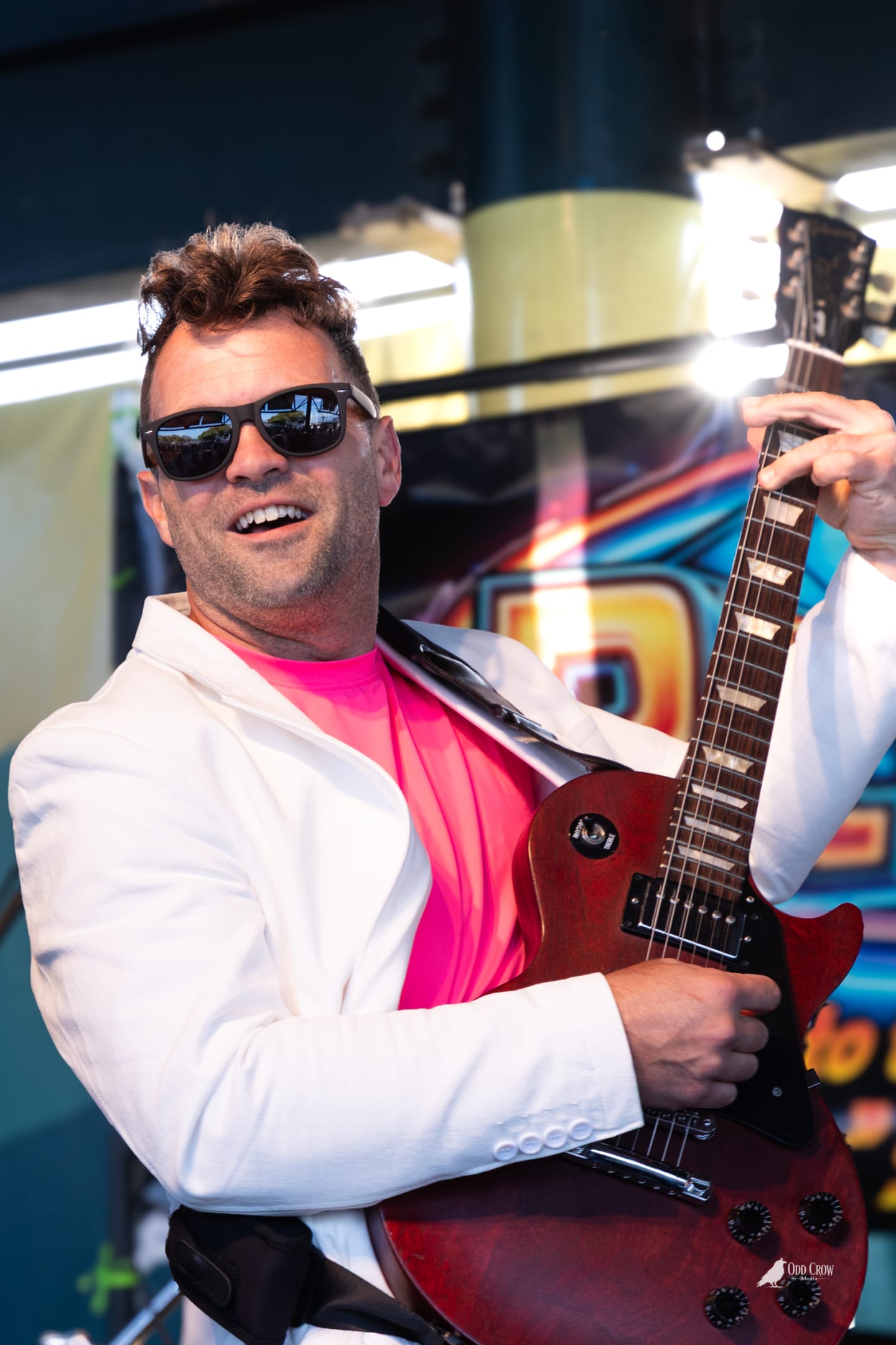 Guitarist in white blazer and hot pink shirt grinning at the camera while holding a red Gibson Les Paul guitar on the Rewind stage
