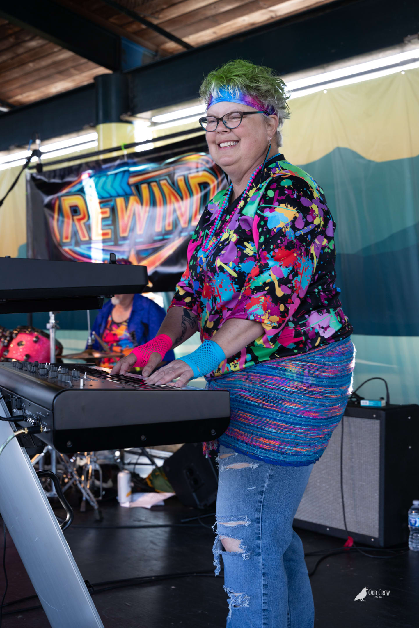 Keyboard player in paint-splatter shirt and green hair smiling broadly behind her keys on the Rewind stage at Concerts on the Pier Port Angeles