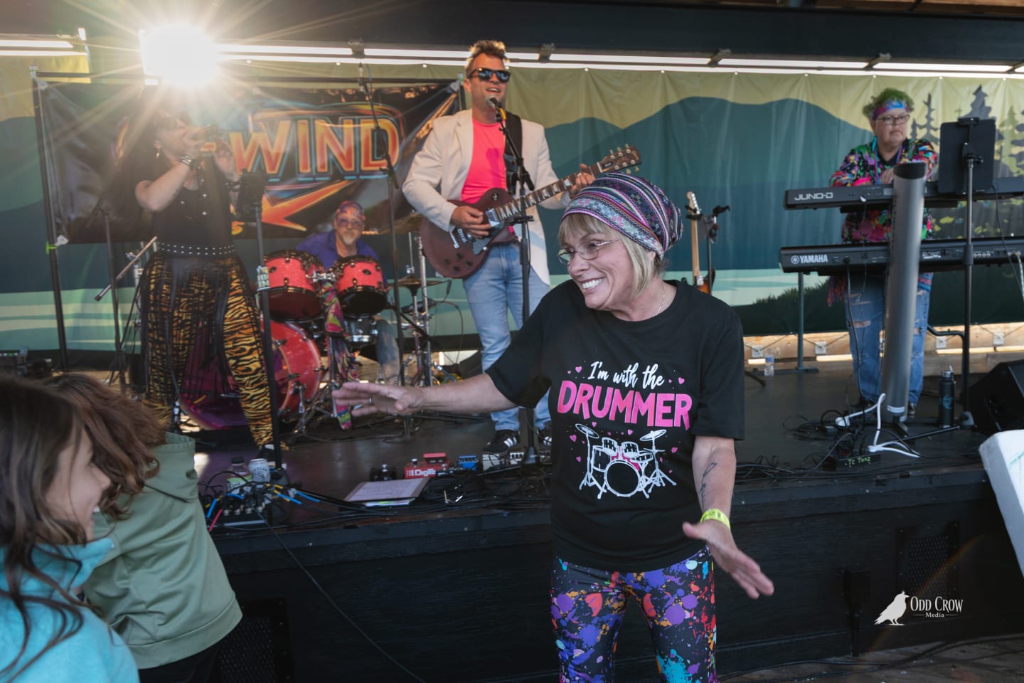 Woman in 'I'm with the Drummer' shirt dancing with a child near the front of the stage at Concerts on the Pier, full band visible performing behind them in late afternoon light