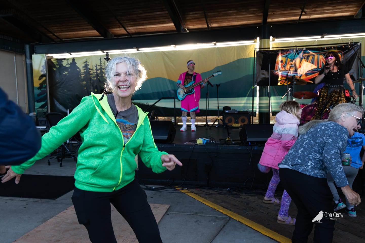 Older woman in bright green zip-up hoodie dancing joyfully at the front of the crowd at Concerts on the Pier, with the full Rewind band performing on stage in the background