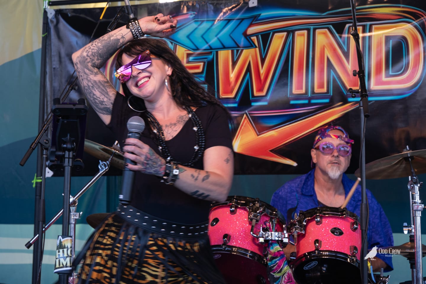 Lead singer of Rewind with arm raised and pink mirrored sunglasses smiling mid-song in front of the bold Rewind Back to the 80s banner, drummer visible behind her