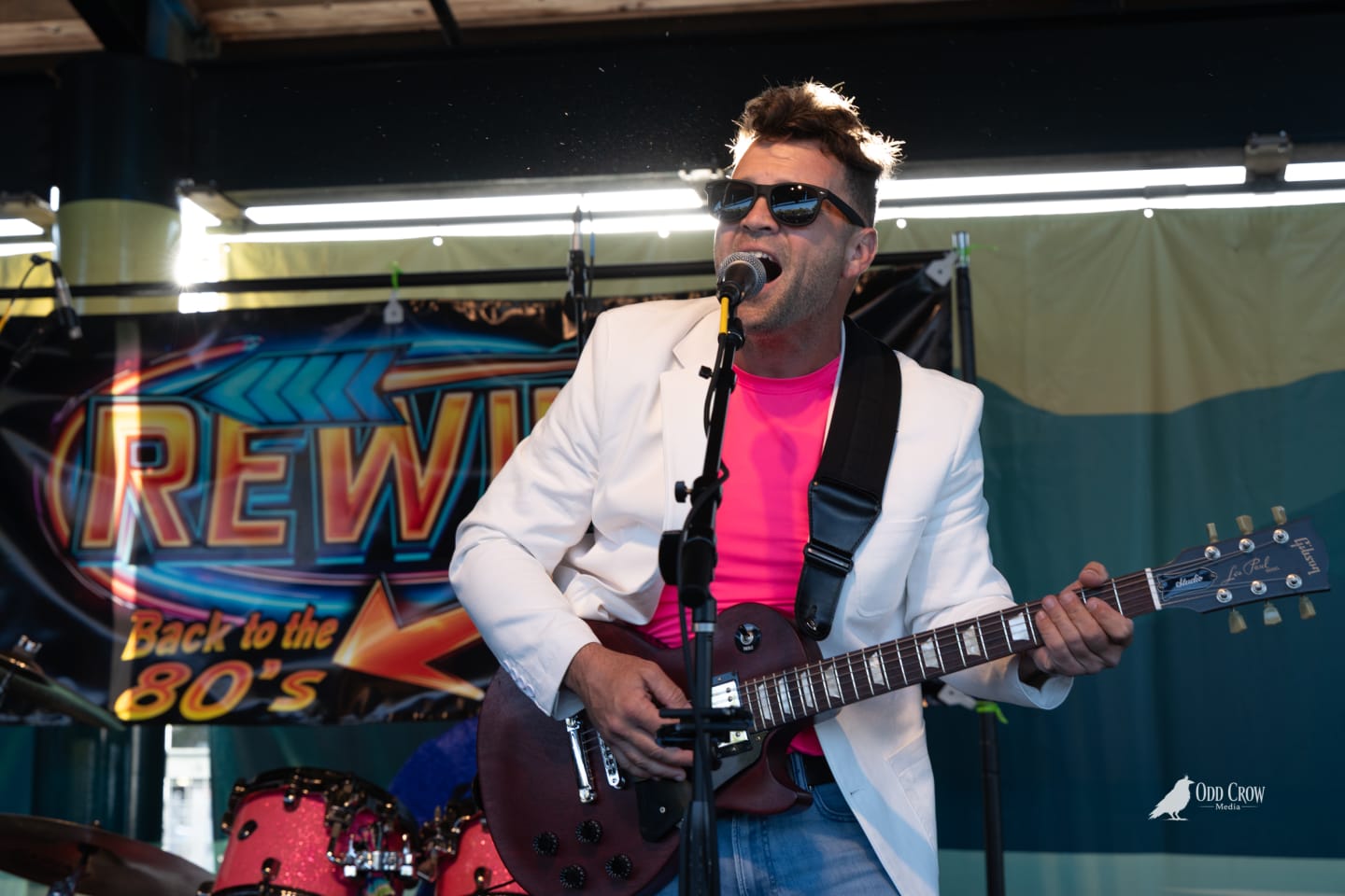 Guitarist and vocalist of Rewind in white blazer and pink shirt singing into a mic while playing red Gibson Les Paul guitar, Rewind Back to the 80s banner behind him