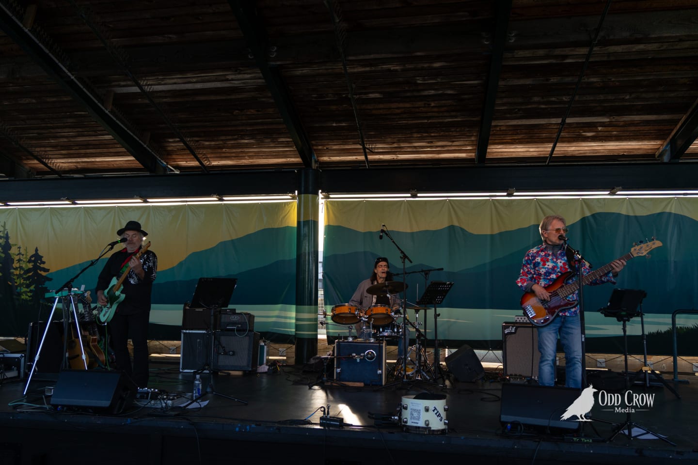 Three-piece band The Foot Stompers performing on the covered pier stage in Port Angeles, with a painted Pacific Northwest mountain and forest backdrop lit by strip lights