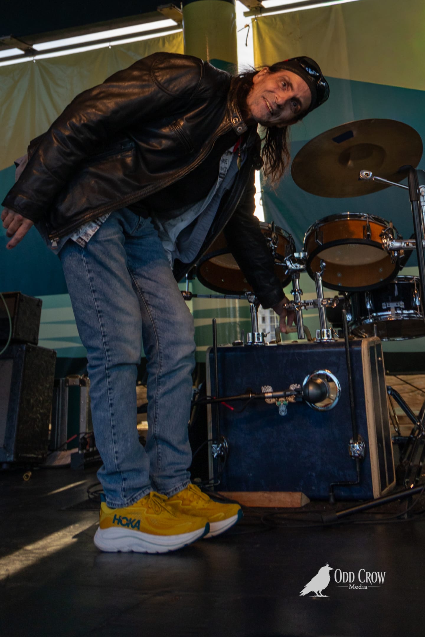 Drummer of the Foot Stompers leaning toward the camera from behind his kit with a grin, wearing a leather jacket and yellow HOKA sneakers on the pier stage