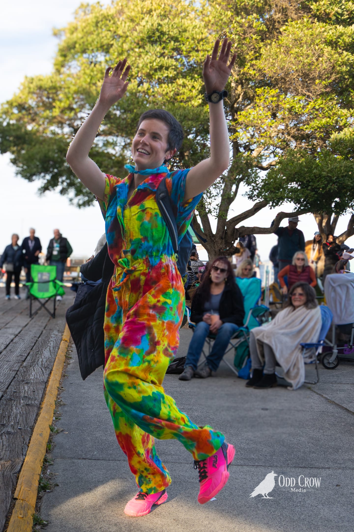 Woman in a full tie-dye jumpsuit dancing with arms raised on the waterfront sidewalk at Concerts on the Pier, audience members seated behind her