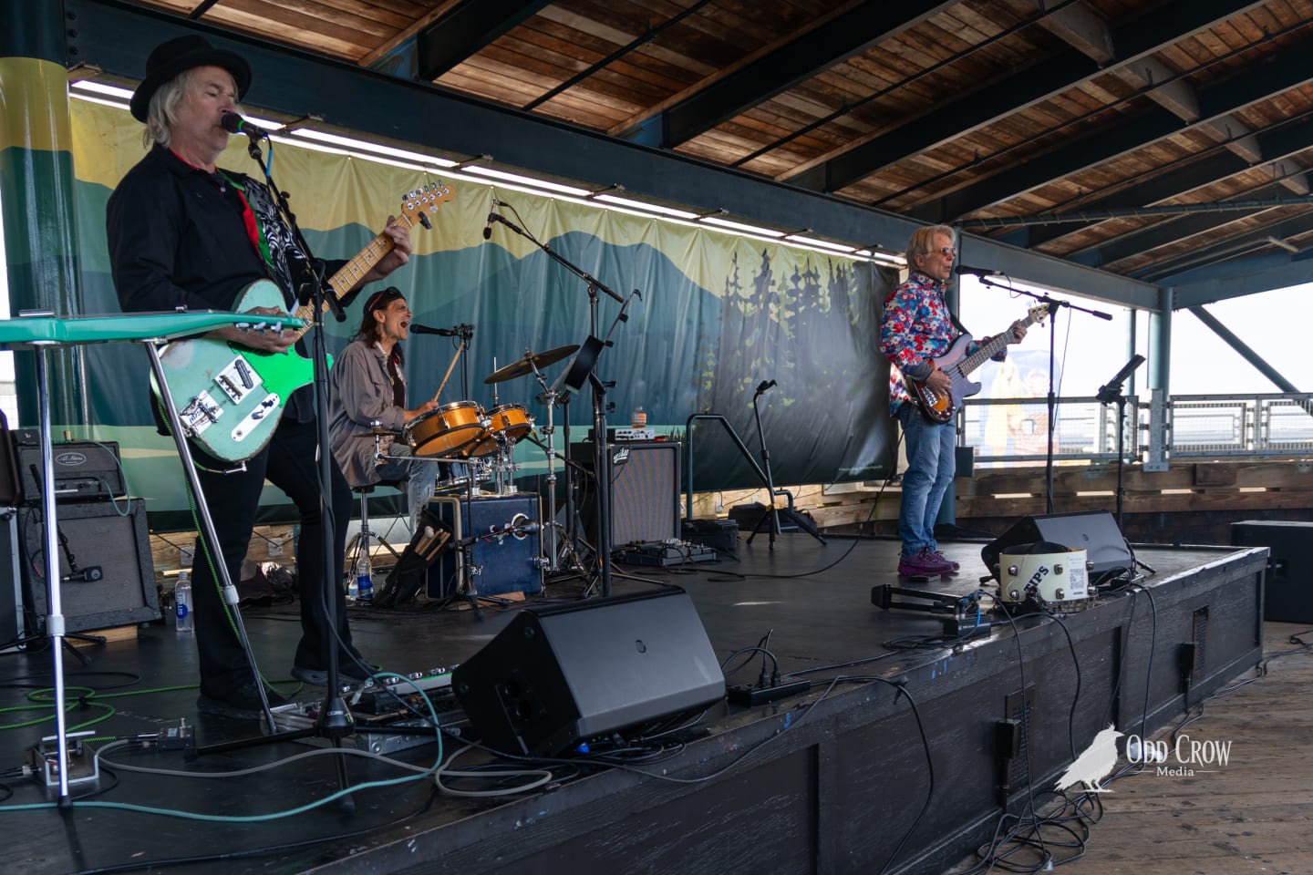 Foot Stompers guitarist and drummer sharing a look between songs, both smiling, acoustic guitar in hand, painted mountain backdrop behind them