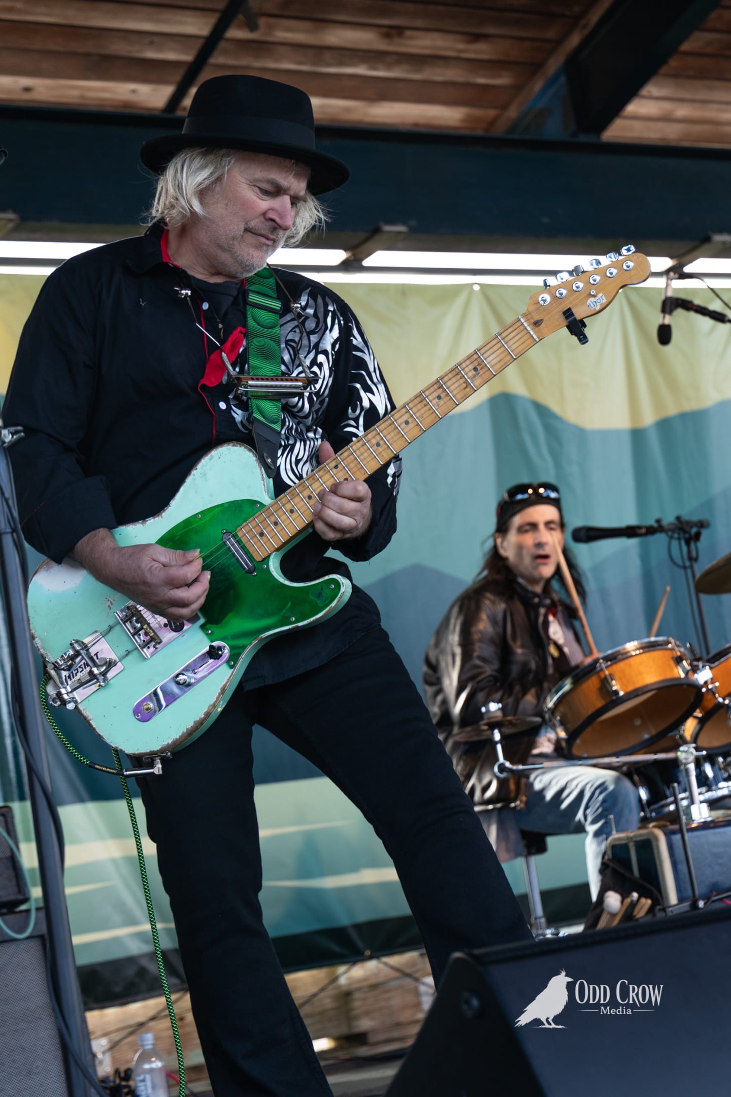 Foot Stompers drummer playing the cymbal with sticks in both hands, big grin, leather jacket, HOKA sneakers visible, custom suitcase bass drum below