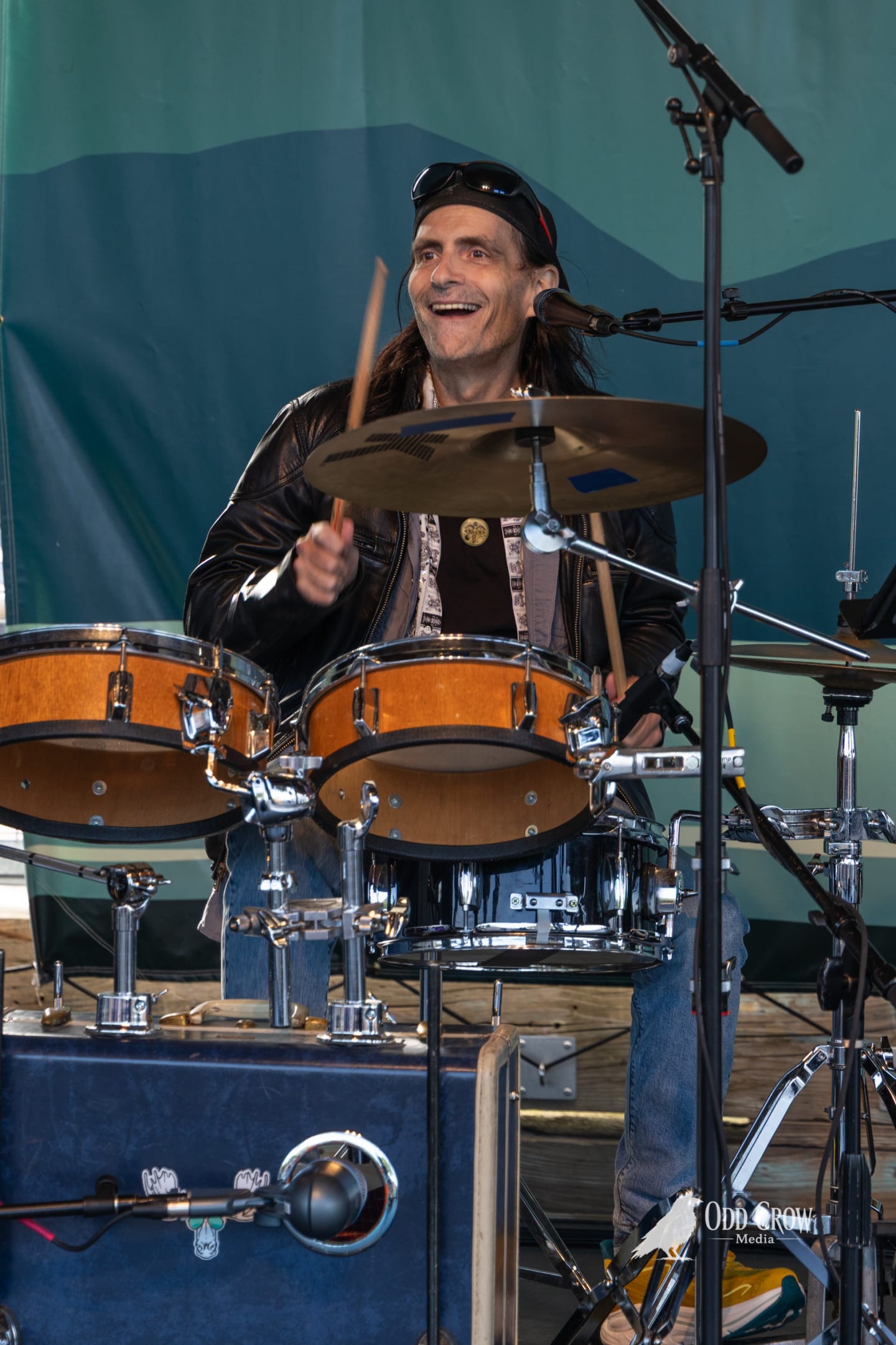 Foot Stompers drummer singing into a mic while playing, framed through the blurred neck of the guitarist's acoustic guitar in the foreground