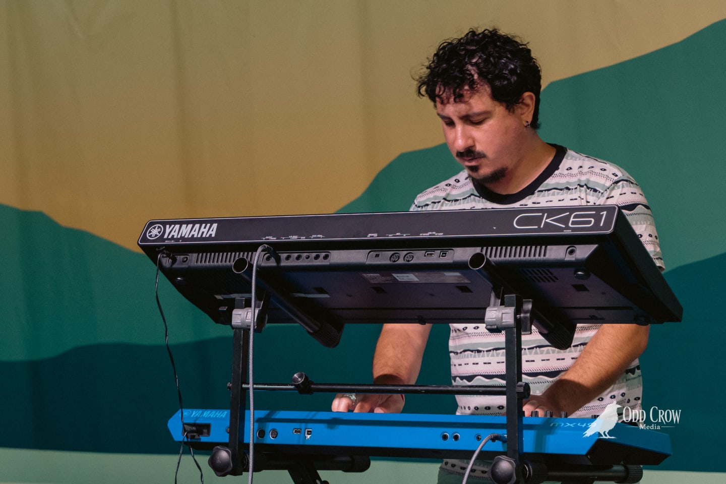Keyboardist playing stacked Yamaha CK61 and MX49 synthesizers on an outdoor stage with colorful backdrop.