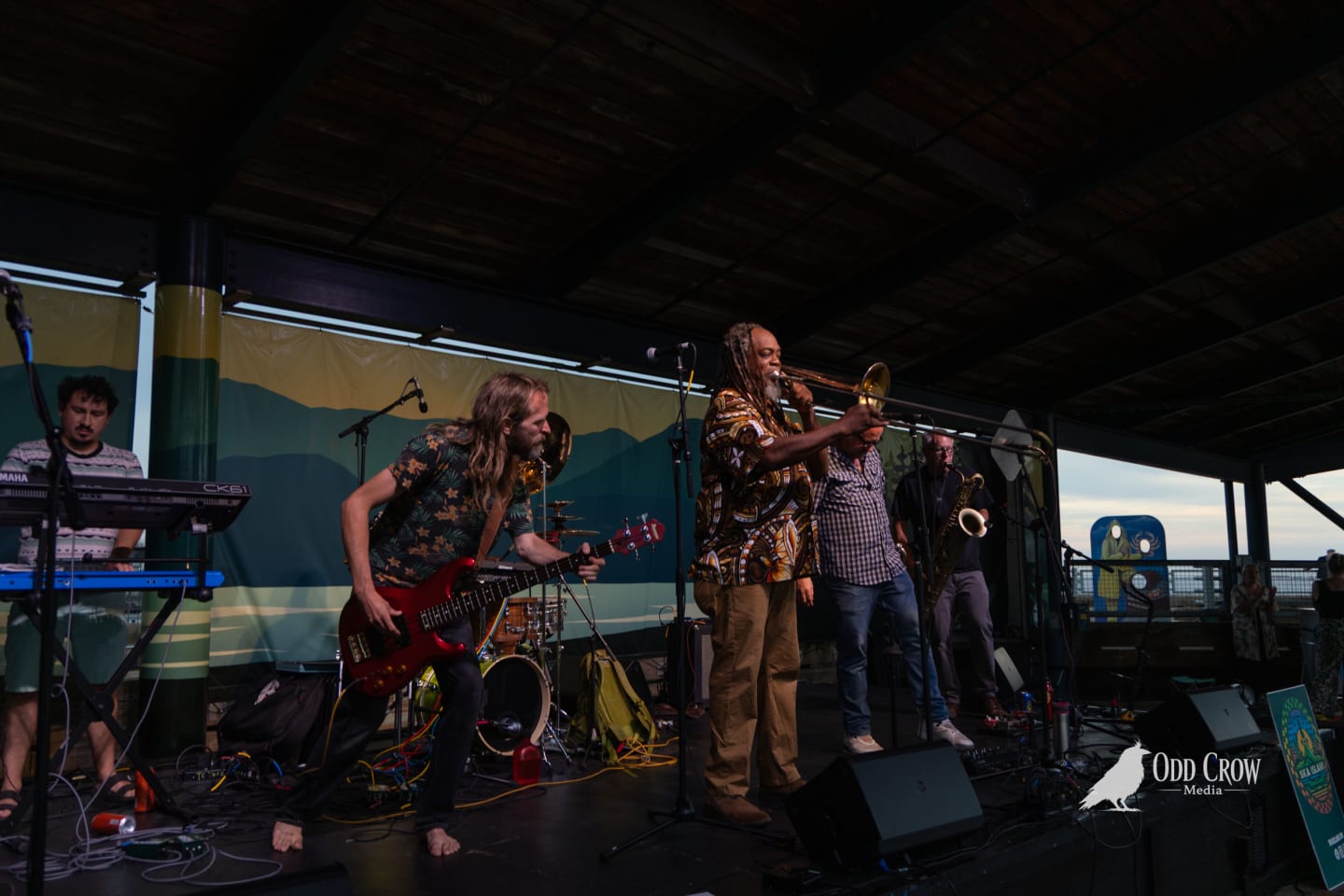 Ska band performs on outdoor covered pier stage, trombonist and guitarist energetically playing at sunset.
