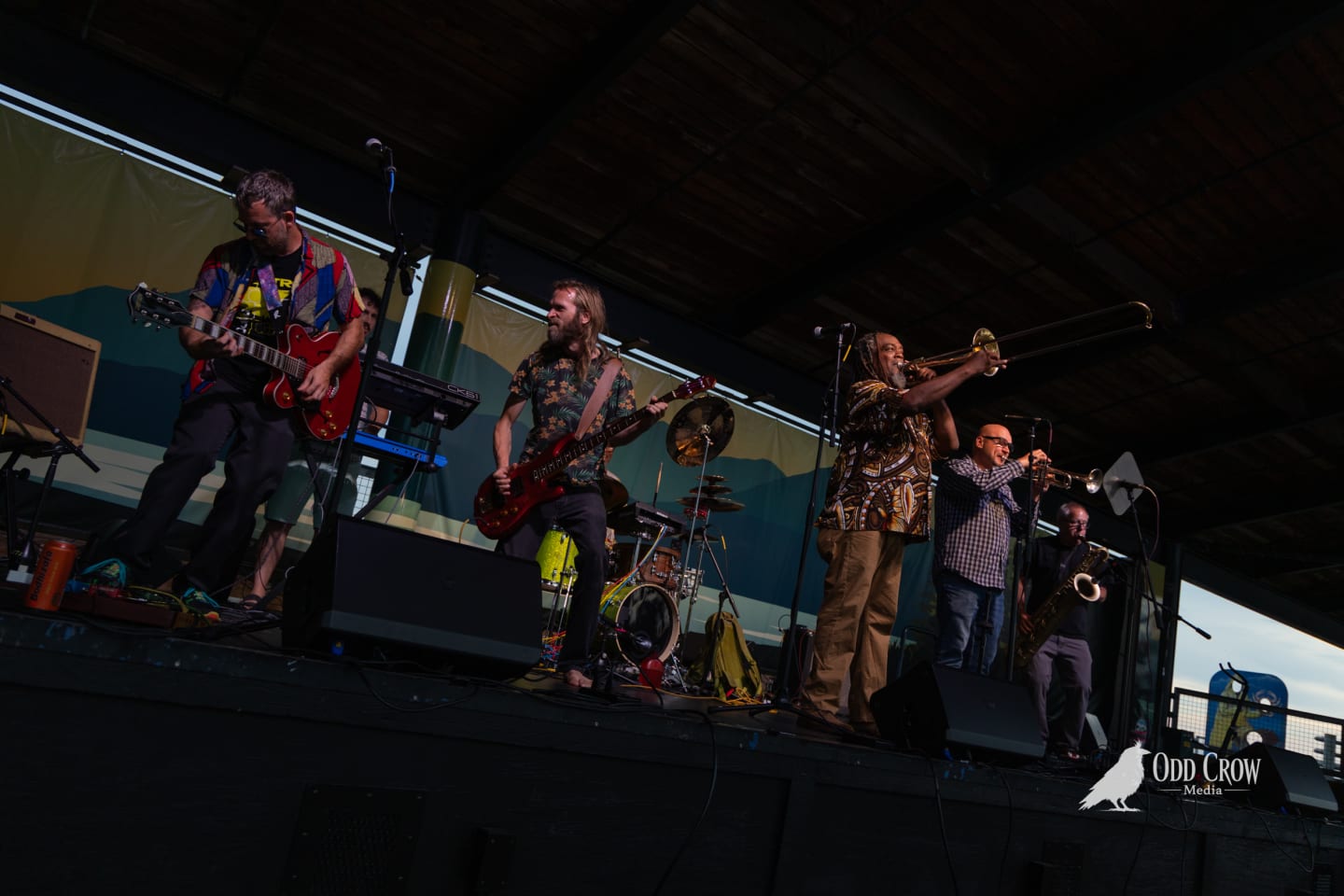 Live band performs on outdoor pier stage with guitars, trombone, trumpet, and saxophone at sunset.