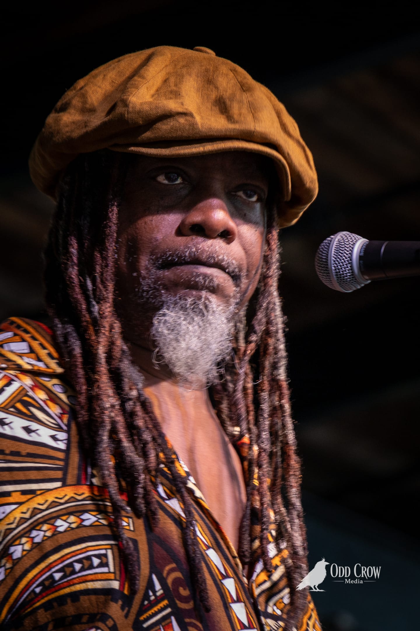 Reggae vocalist with dreadlocks, gold flat cap, and African-print shirt performing at a microphone onstage.