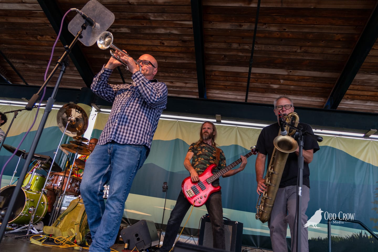 Trumpet and baritone saxophone players perform alongside a bassist on a covered outdoor festival stage.
