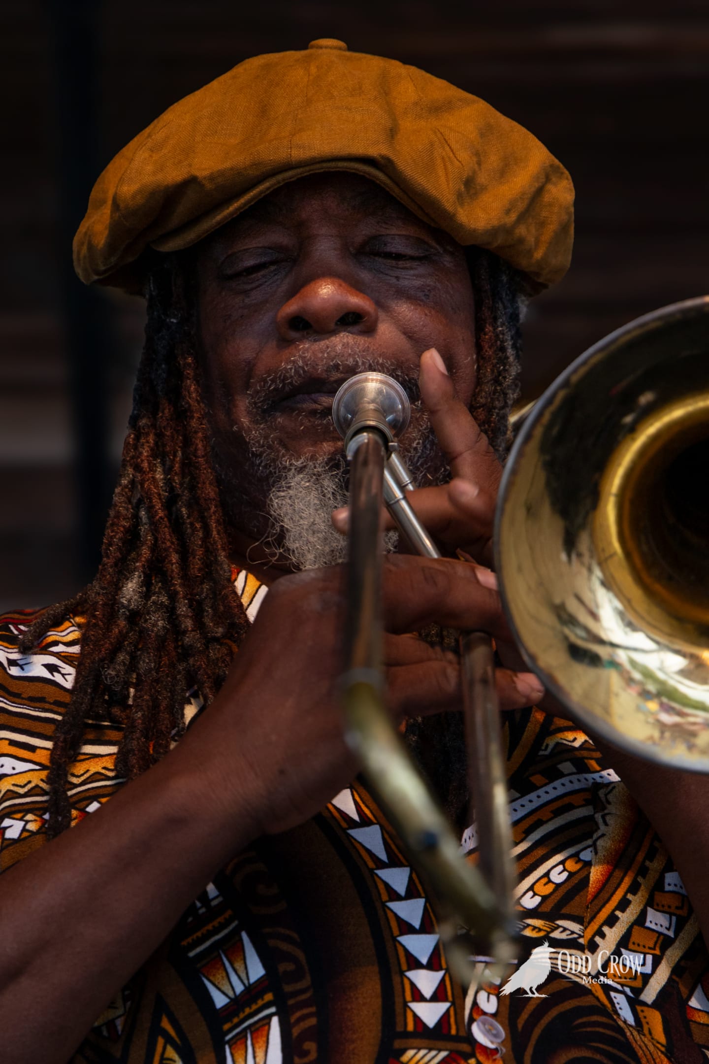 Older man with dreadlocks and gold flat cap plays trombone, eyes closed, in patterned African-print shirt.