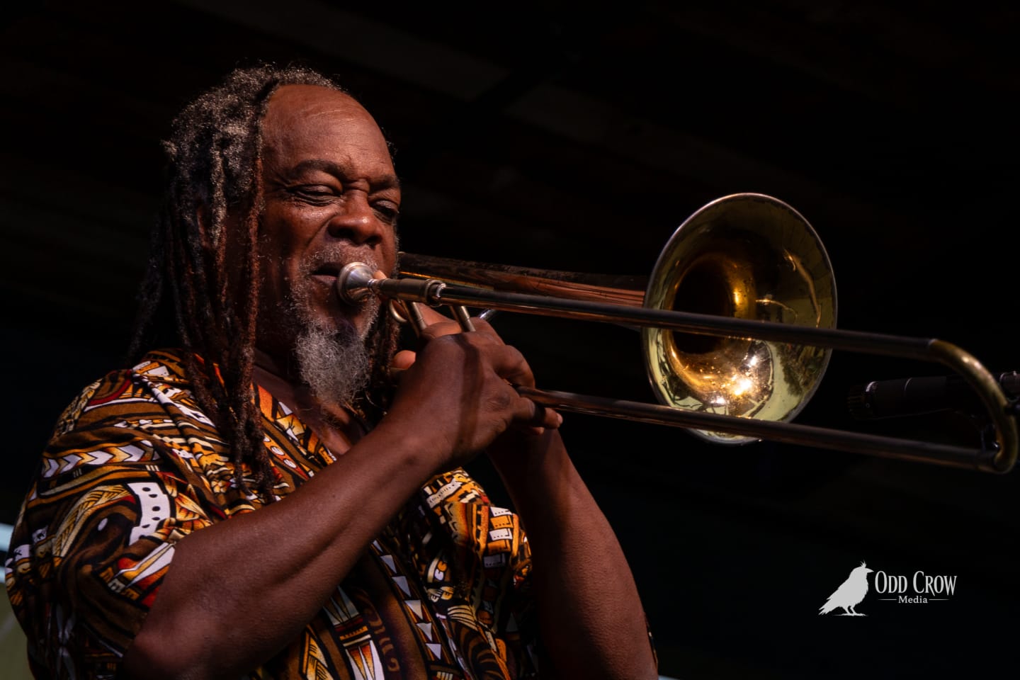 Dreadlocked trombonist plays passionately on outdoor stage, wearing a colorful African-print shirt.