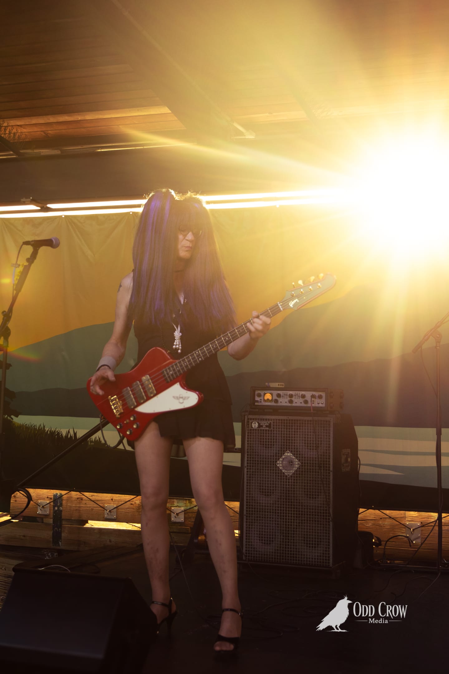 Guitarist in black dress and long dark hair plays red electric guitar on sunlit outdoor stage.