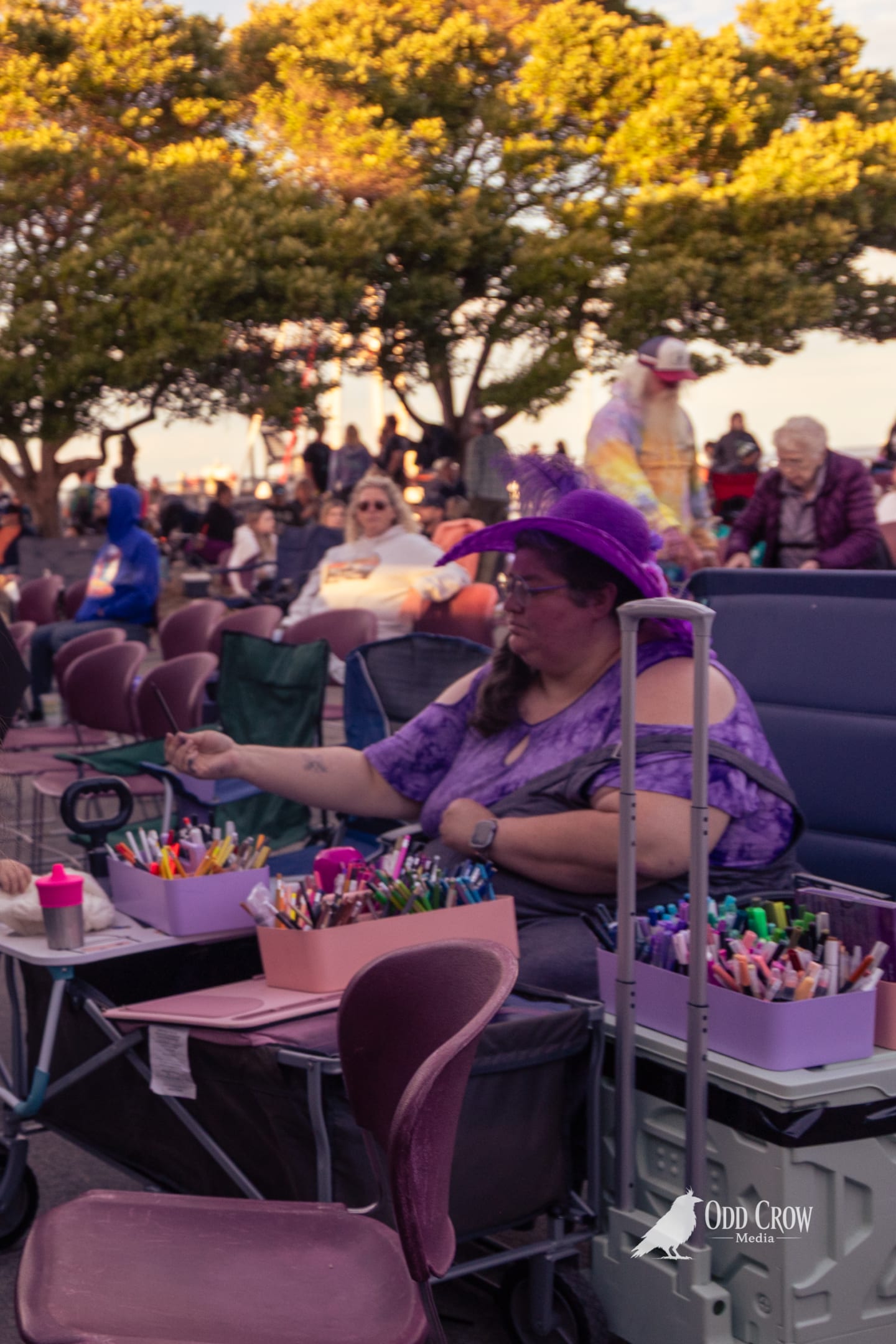 Woman in purple hat and outfit sorting markers at an outdoor waterfront concert.