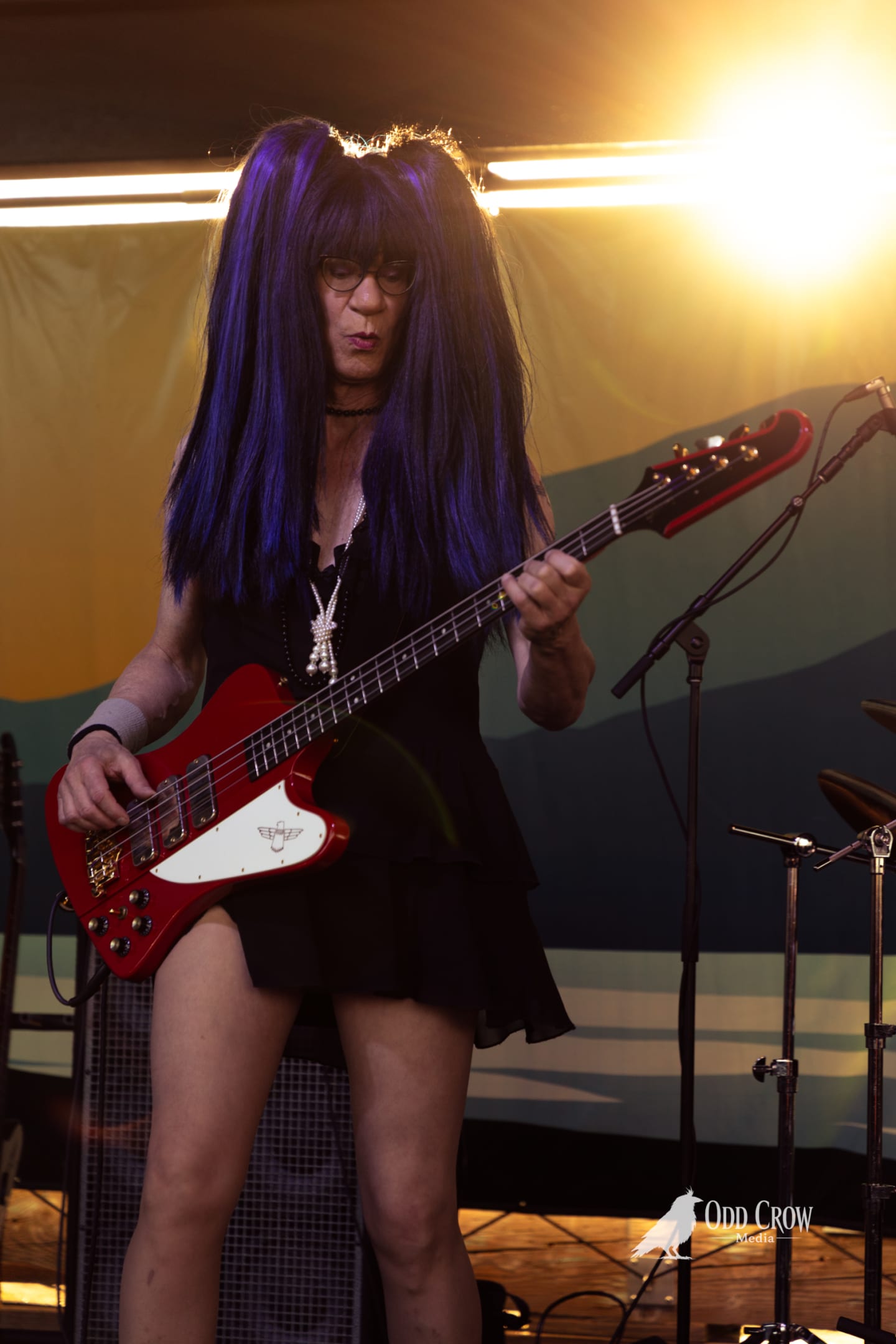 Bassist with long purple hair plays a red Thunderbird bass on an outdoor stage under warm stage lighting.