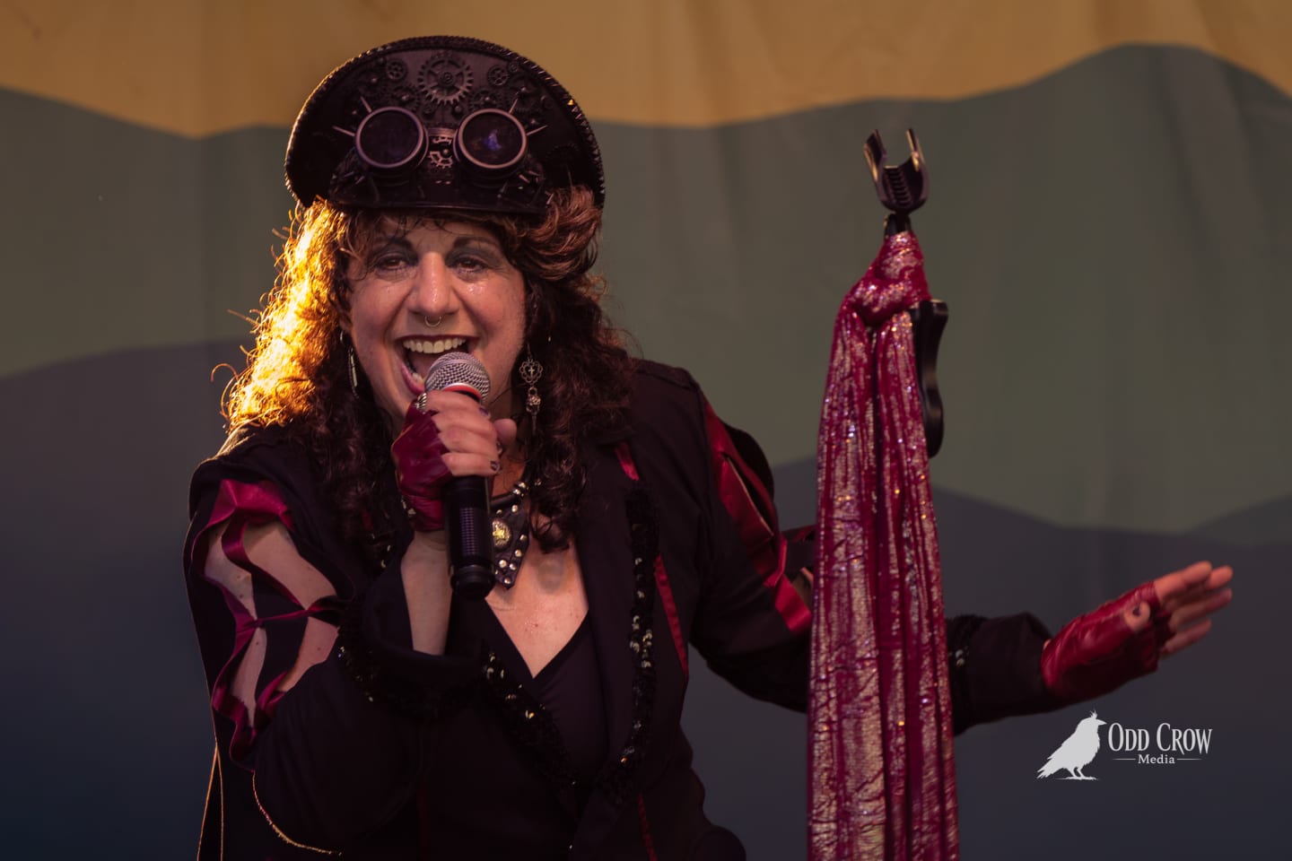 Steampunk-costumed performer sings into microphone on stage, wearing gear-adorned top hat with goggles.