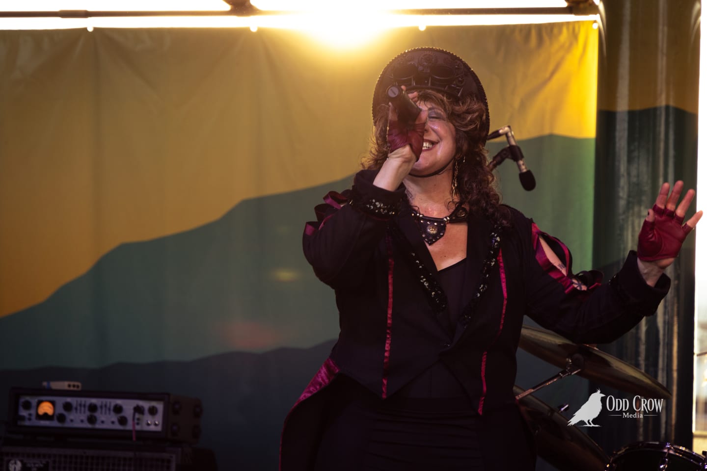Steampunk-costumed vocalist singing into microphone on stage, arms raised, backlit by warm stage lighting.