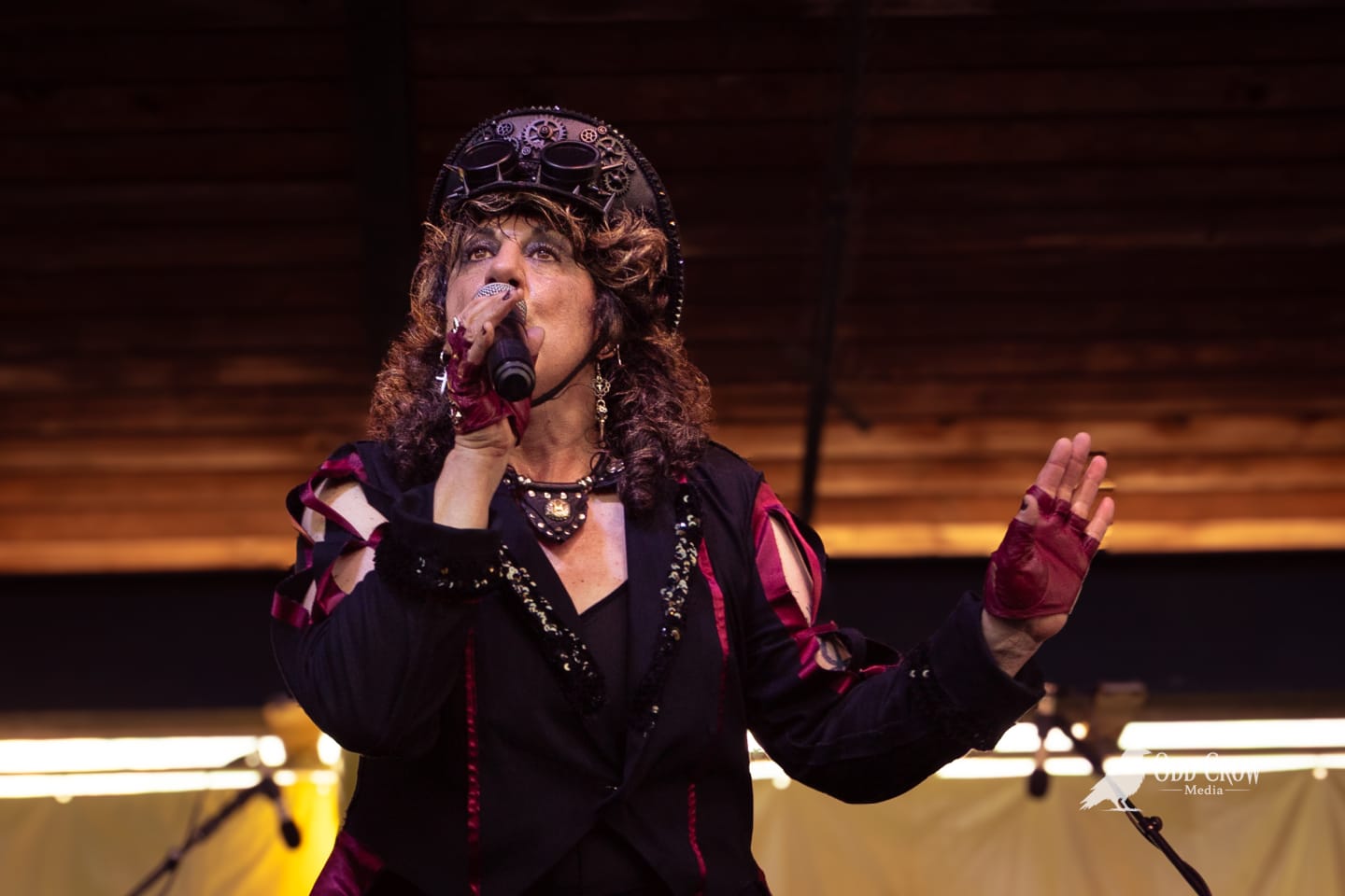 Female steampunk vocalist in geared hat and fingerless gloves performing on stage with handheld microphone.