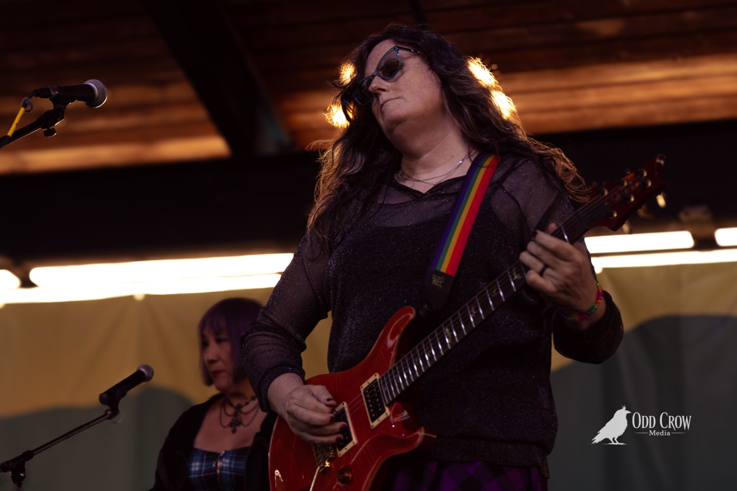 Guitarist in sunglasses plays red electric guitar with rainbow strap on an outdoor stage.