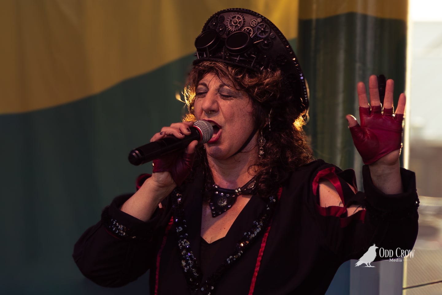 Steampunk-costumed vocalist singing passionately into microphone, one fingerless-gloved hand raised, performing live on stage.