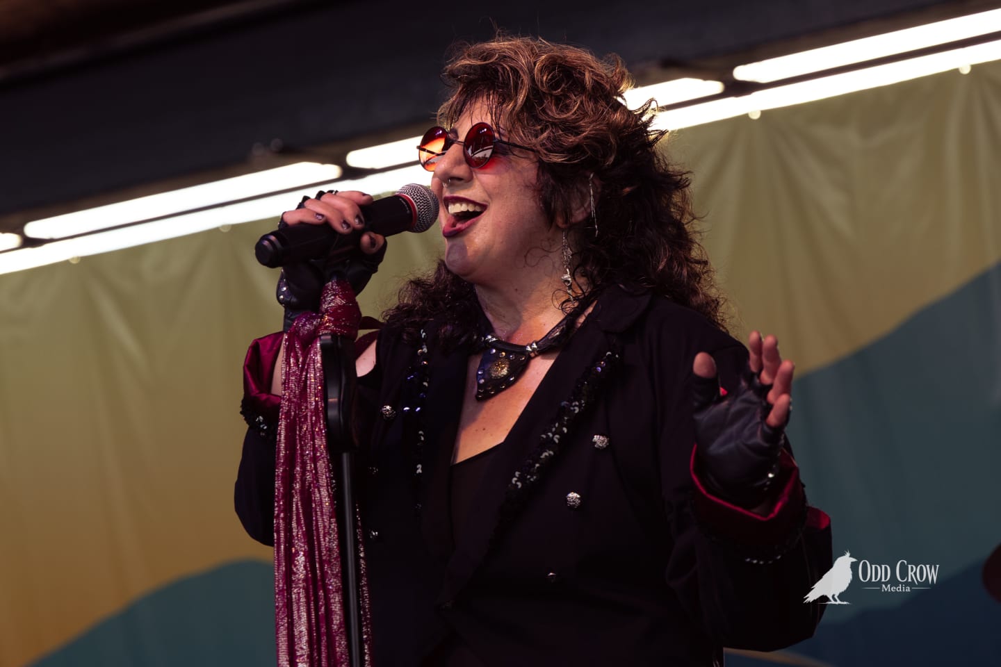 Curly-haired female vocalist in black sequined jacket and red sunglasses performs energetically on outdoor stage.
