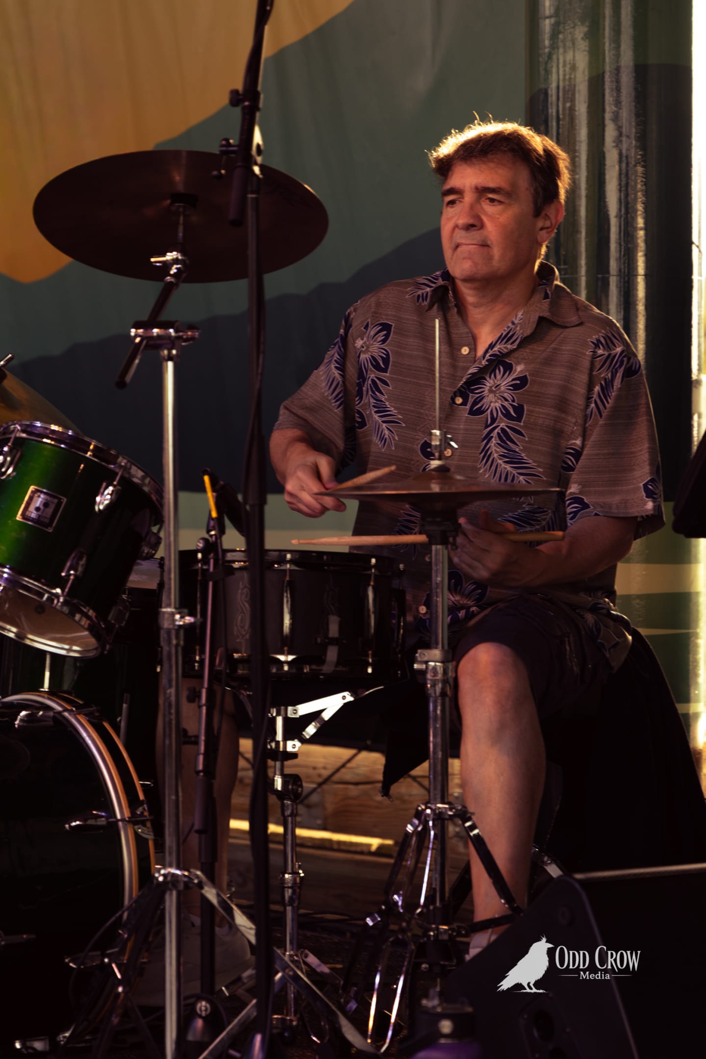 Drummer in a floral shirt plays cymbals on an outdoor stage at a pier concert.