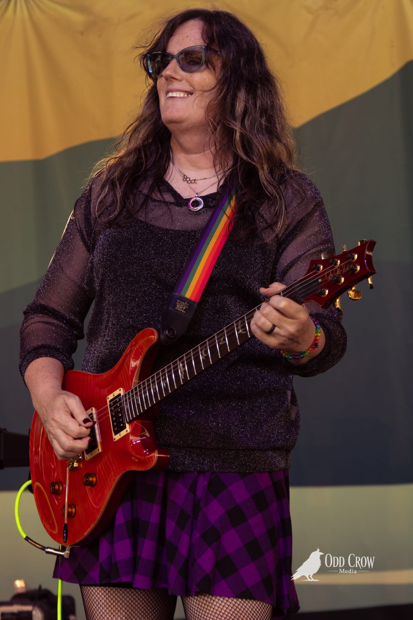 Smiling guitarist plays red PRS electric guitar with rainbow strap on outdoor festival stage.