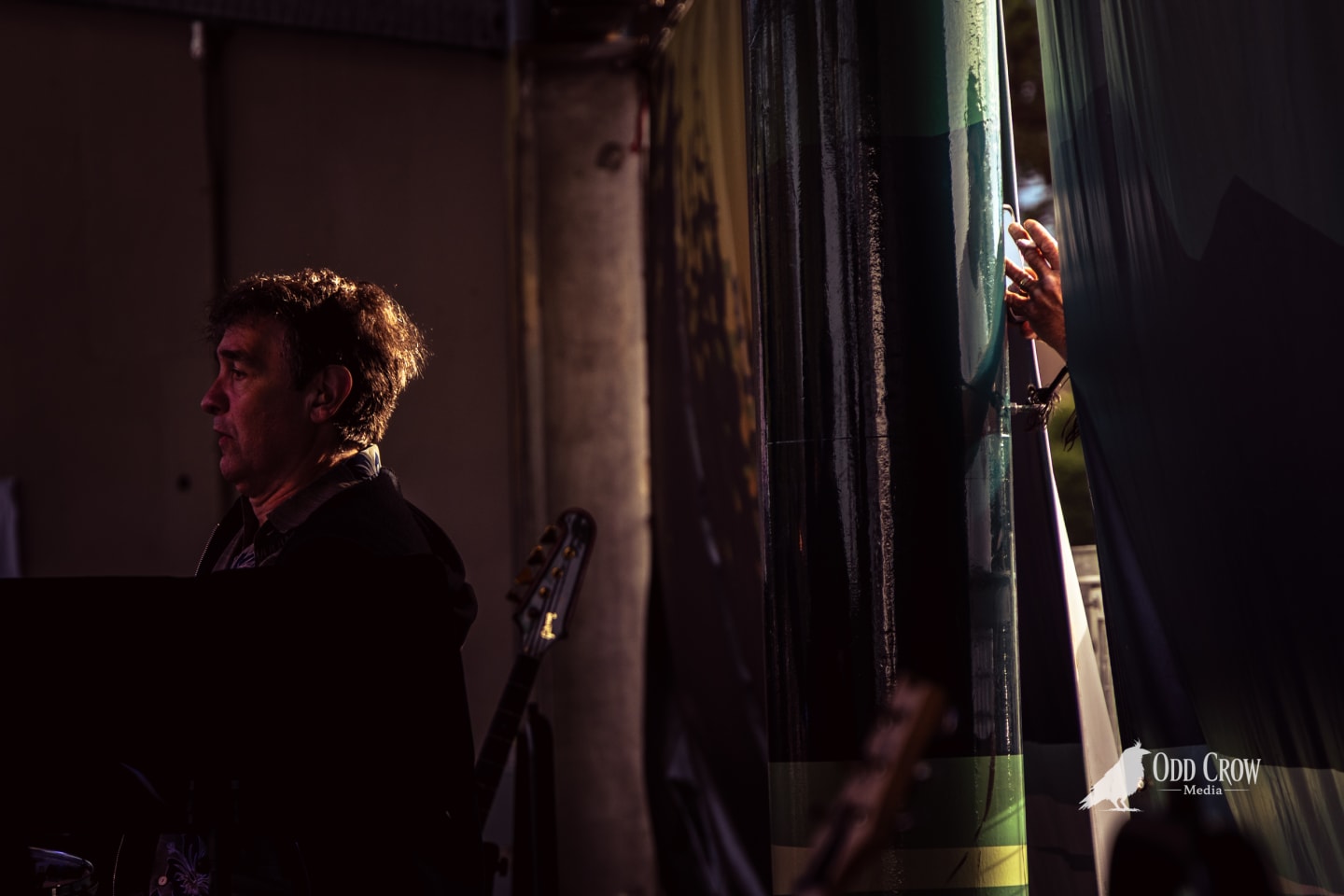 Musician stands in profile backstage, lit by warm stage light, guitar visible behind him.