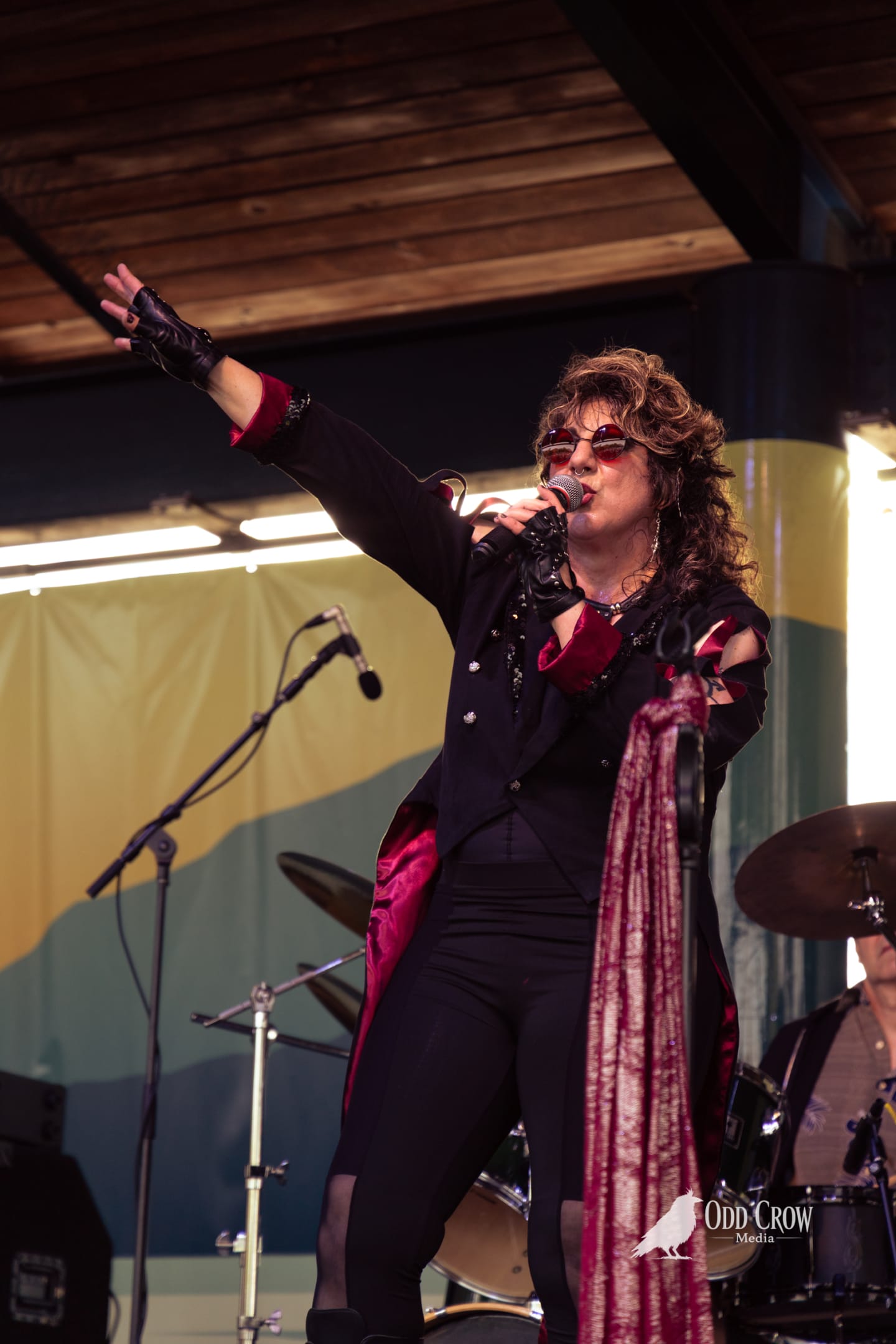 Rock vocalist in dramatic black and crimson coat performs on stage, arm raised, singing into microphone.