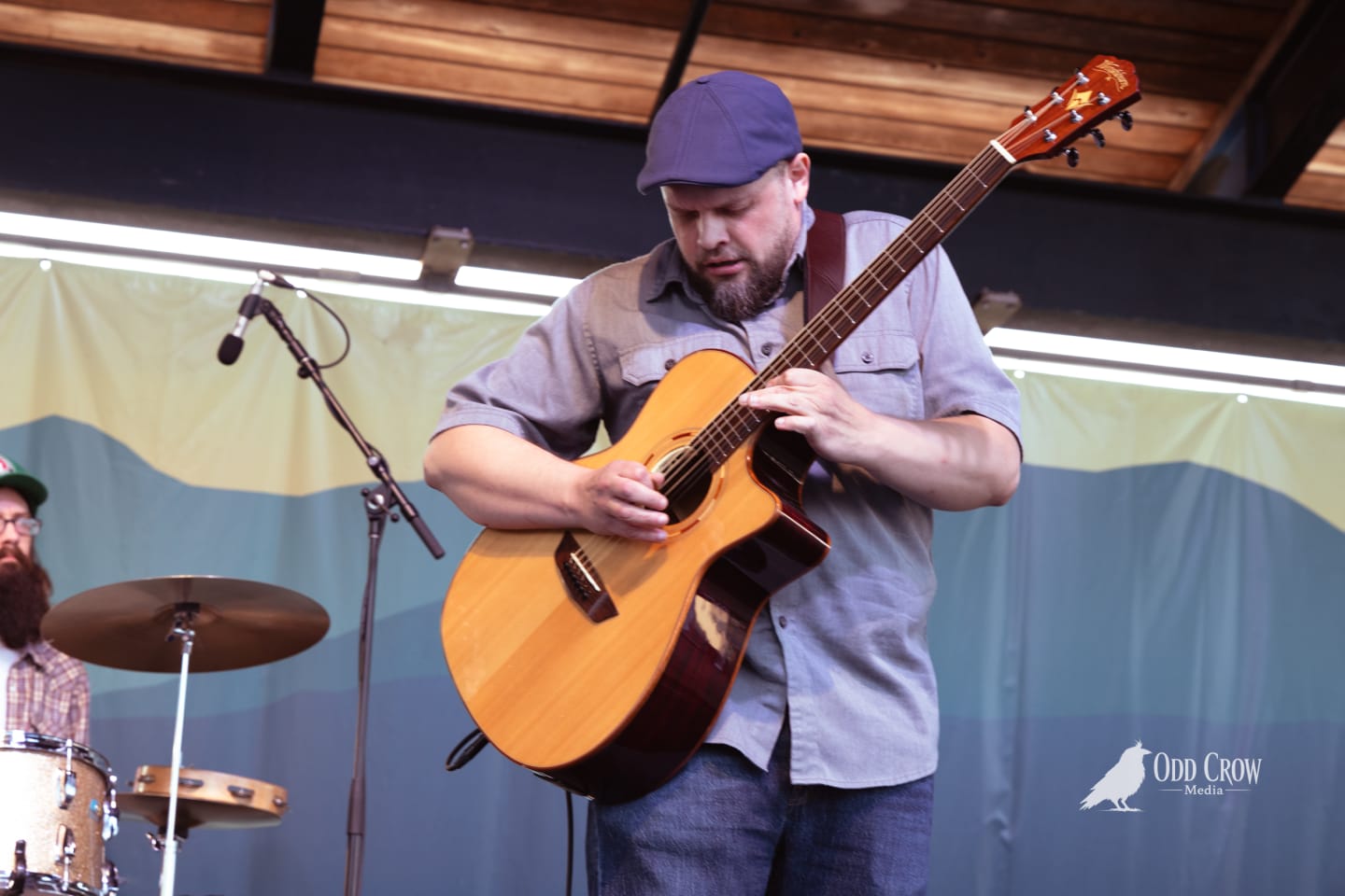 Bearded guitarist in blue cap plays acoustic guitar on outdoor stage with drummer visible behind him.