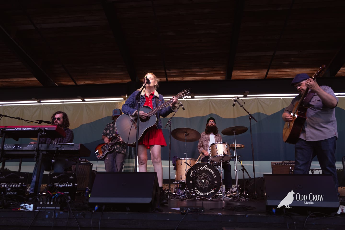 Female vocalist plays acoustic guitar on outdoor stage with full band including keys, guitar, and drums.