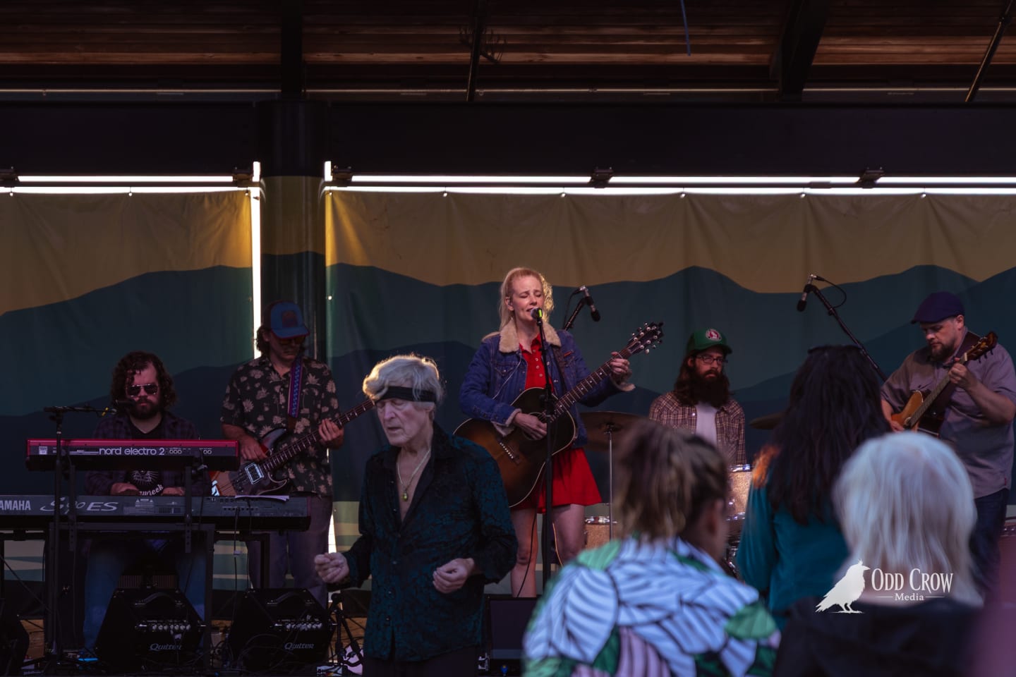 Band performs on outdoor stage beneath mountain backdrop mural, female vocalist strums acoustic guitar at microphone.