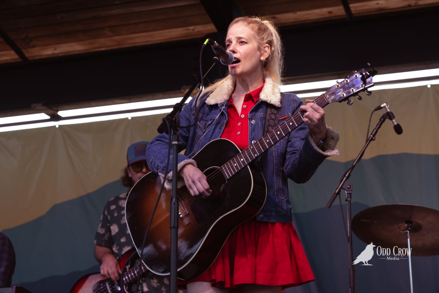 Blonde woman in red dress and denim jacket sings and plays acoustic guitar on outdoor stage.